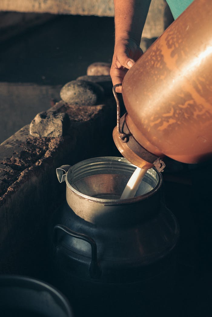 A hand pouring milk into a stainless steel container, captured in a warm, rustic setting.